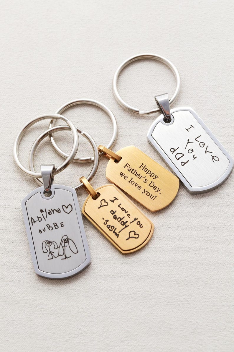 May include: Three metal dog tag keychains with split rings. One silver tag features a child's drawing and the text 'Adilone BUBBE'. One gold tag reads 'Happy Father's Day, we love you!' and 'I love you daddy -Sasha'. One silver tag reads 'I Love You dad'.