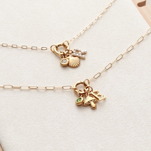 May include: Two gold necklaces with charm pendants. One necklace features a seashell, a diamond, and a letter charm. The other necklace has a mushroom, a green gem, and a letter charm. Both necklaces have delicate chain links.