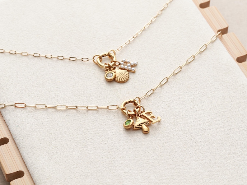 May include: Two gold necklaces with charm pendants. One necklace features a seashell, a diamond, and a letter charm. The other necklace has a mushroom, a green gem, and a letter charm. Both necklaces have delicate chain links.