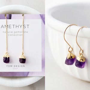 May include: Amethyst gemstone earrings with gold accents. The earrings are on a white surface and have a delicate chain design. The text "Amethyst natural gemstone collection TOM DESIGN SHOP" is on a card behind the earrings.