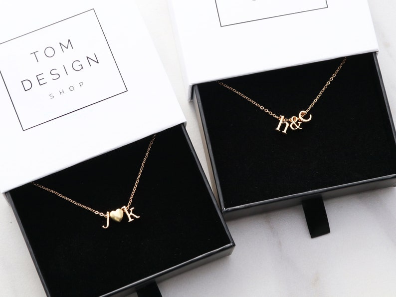 May include: Two gold necklaces in black boxes. One necklace has a heart charm and the letters "J" and "K". The other necklace has the letters "H" and "C". The boxes have the text "TOM DESIGN SHOP" on the top.