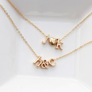 May include: Two gold-toned necklaces with delicate chains. The top necklace features the letters J and K separated by a small heart charm. The bottom necklace displays the lowercase letters h and c connected by an ampersand symbol.