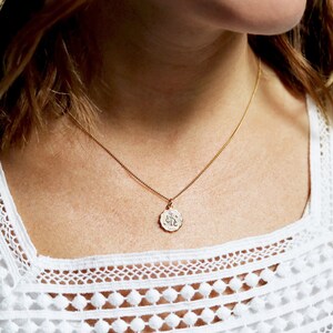 May include: A gold chain necklace with a small, round pendant featuring a delicate design. The pendant is worn by a person with long, brown hair and a white lace top.