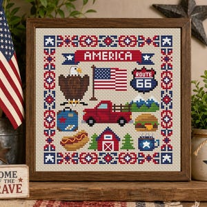 May include: A framed cross-stitch sampler featuring the text AMERICA, a bald eagle, a US flag, a Route 66 sign, a red pickup truck, mountains, a suitcase, a hot dog, a barn, a pine tree, a burger, and a blue mug with a white star, all in a geometric border.