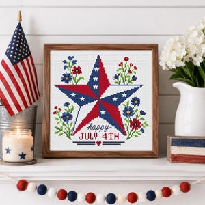 May include: A framed cross-stitch design featuring a red, white, and blue star with the text "happy JULY 4TH." The design is surrounded by floral accents and is displayed on a white shelf with an American flag, candle, and decorative garland.