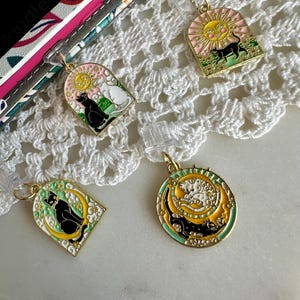 May include: Four gold-toned cat-themed charms with various designs. One charm features a black cat and a white cat under a pink sun. Another shows a black cat under a smiling sun. The other two charms feature a black cat on a crescent moon.