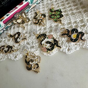 May include: A collection of gold-toned charms with black and white snake designs, some adorned with floral and celestial accents. The charms are displayed on a white lace surface, showcasing their intricate details and varied patterns.