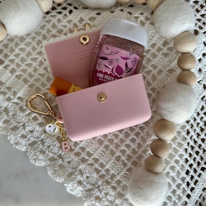 May include: A light pink cardholder and matching case with a gold snap closure. The case holds a small bottle of hand sanitizer and a lip balm. A gold keychain with a charm and the letter "B" hangs from the case.