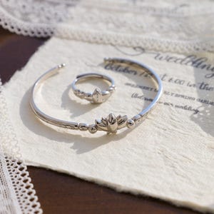 Könnte beinhalten: Ein silberfarbenes Armband- und Ring-Set. Das Armband hat ein Blumendesign mit Perlenakzenten. Der Ring spiegelt das Blumenmotiv wider. Der Schmuck liegt auf strukturiertem Papier, mit einer Hochzeitseinladung im Hintergrund.
