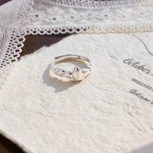 Könnte beinhalten: Ein verstellbarer Silberring mit einem blattförmigen Design. Der Ring hat ein geflochtenes Band und ein zentrales Blattmotiv mit drei kleinen dekorativen Punkten. Der Ring wird auf einer strukturierten, cremefarbenen Oberfläche präsentiert.