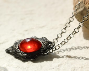 May include: A silver necklace with a red gemstone pendant. The pendant is shaped like a stylized eye with intricate details.