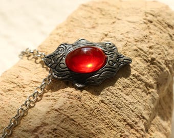 May include: A silver chain necklace with a red gemstone pendant. The pendant is an ornate design with a swirling pattern.