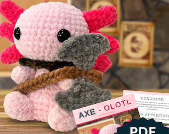 May include: Pink and red crocheted axolotl plush toy holding a gray crocheted axe. The toy is sitting on a wooden surface. The text "AXE - OLOTL Crochet Pattern" is visible on a white card behind the toy.