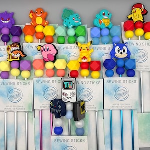 May include: A collection of sewing sticks featuring various cartoon character designs. Each stick has a colorful bead and geometric shape design, with characters like Pikachu, Sonic, and Kirby. The packaging reads "SEWING STICKS" and "HOLDS TOGETHER CROCHETED PLUSHIES."