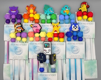 May include: A collection of sewing sticks featuring various cartoon character designs. Each stick has a colorful bead and geometric shape design, with characters like Pikachu, Sonic, and Kirby. The packaging reads "SEWING STICKS" and "HOLDS TOGETHER CROCHETED PLUSHIES."