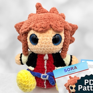 May include: A crocheted doll of a character with spiky brown hair, large blue eyes, and a red and black outfit. The doll is seated with yellow shoes and a blue belt. A card with the text "SORA Crochet Pattern" and "PDF Pattern" is in the foreground.