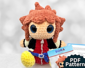 May include: A handmade crochet doll of a character with spiky brown hair, large blue eyes, and a red and black outfit. The doll is seated with yellow shoes and a blue belt. A card with the text "SORA Crochet Pattern" and "PDF Pattern" is in the foreground.