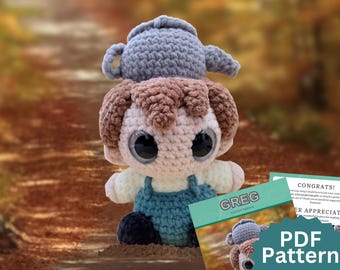 May include: A crochet doll with brown hair, large black eyes, and a light skin tone, wearing blue overalls over a white shirt. A gray crocheted teapot sits on the doll's head. A card labeled GREG Crochet Pattern is visible in the foreground.