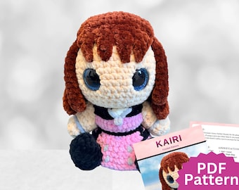 May include: A crocheted plush doll with brown yarn hair, large dark blue eyes, and a cream-colored face. The doll wears a black and pink outfit with light blue accents. Beside the doll is a document titled KAIRI Crochet Pattern PDF Pattern.