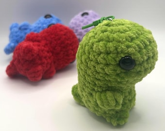May include: A close-up of a green crocheted dinosaur keychain with a black eye. Other crocheted dinosaurs in red, blue, and purple are in the background. The keychains are made of a soft, textured yarn.
