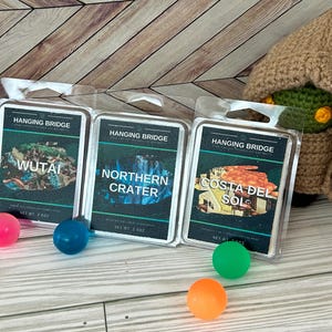 May include: Three Hanging Bridge wax melt packages with scenic images of Wutai, Northern Crater, and Costa del Sol. A crocheted figure in a brown hooded robe is on the right. Colorful bouncy balls are scattered in the foreground.