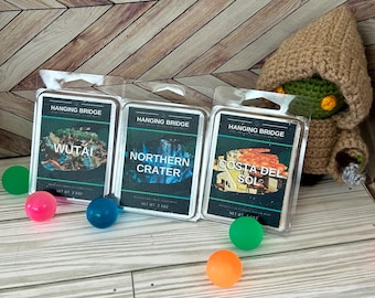 May include: Three Hanging Bridge wax melt packages with scenic images of Wutai, Northern Crater, and Costa del Sol. A crocheted figure in a brown hooded robe is on the right. Colorful bouncy balls are scattered in the foreground.