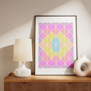 Puede incluir: Una impresión enmarcada con un patrón geométrico de triángulos rosas, amarillos, azules y blancos. La impresión está colgada en una pared sobre una cómoda de madera con una lámpara blanca y un jarrón de cerámica blanco.