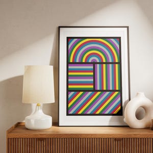 以下が含まれることがあります： 虹、縞模様、斜めの線で構成されたカラフルな幾何学模様の額装されたプリント。プリントは、白いランプと白いセラミックの花瓶のある木製の棚に飾られています。