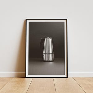 May include: A silver moka pot with a black handle sits on a dark gray surface. The moka pot is a type of stovetop coffee maker.