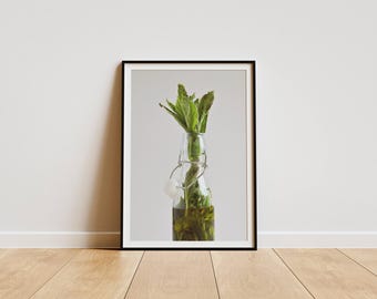 Op de afbeelding: Een ingelijste print van een heldere glazen fles gevuld met groene, bladerige kruiden. De fles heeft een witte stop en staat tegen een witte achtergrond.