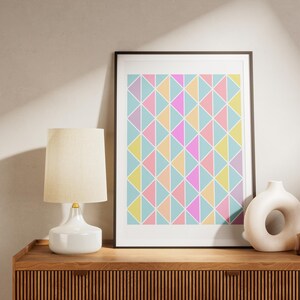 Puede incluir: Una impresión enmarcada con un patrón geométrico de triángulos de color rosa pastel, amarillo y azul. La impresión está colgada en una pared beige claro sobre una cómoda de madera con una lámpara blanca y un jarrón de cerámica blanco.
