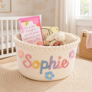 May include: A round, off-white woven cotton rope basket featuring the name Sophie in multi-colored yarn lettering. The basket is decorated with small, crocheted flowers in pink, purple, and blue. Inside are a pink greeting card and a plush toy.