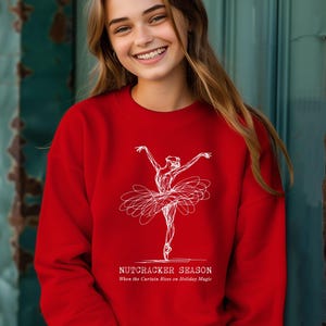 May include: A red sweatshirt with a white sketch of a ballerina. The text "NUTCRACKER SEASON" and "When the Curtain Rises on Holiday Magic" are printed below the ballerina. This festive sweatshirt is perfect for the holiday season.