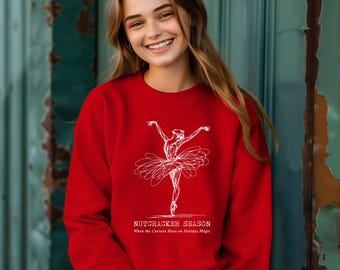 May include: A red sweatshirt featuring a white sketch of a ballerina in a tutu. The text "NUTCRACKER SEASON" and "When the Curtain Rises on Holiday Magic" are printed below the ballerina. The sweatshirt is a festive holiday apparel item.