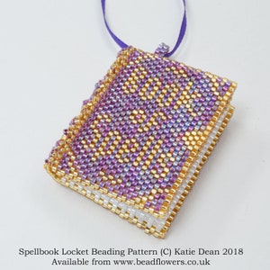 Peut inclure: Un petit médaillon en forme de livre, en perles violettes et dorées. Le médaillon est suspendu à un cordon violet. Le texte "Spellbook Locket Beading Pattern (C) Katie Dean 2018 Available from www.beadflowers.co.uk" est imprimé sous le médaillon.
