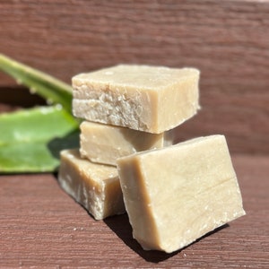 May include: Four bars of natural soap stacked on top of each other. The soap is a light beige colour and has a rough texture. The soap is sitting on a wooden surface.