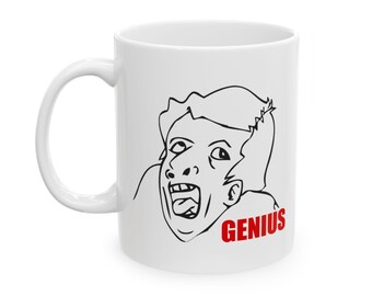 Può includere: Tazza in ceramica bianca con un disegno a linea nera di una persona con la bocca aperta e la lingua fuori. Il testo "GENIUS" è stampato in rosso sotto il disegno.
