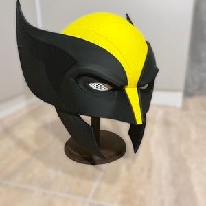 Pode incluir: Uma máscara de Wolverine preta e amarela, exibida em um suporte de madeira. A máscara apresenta uma parte superior amarela e detalhes em preto, incluindo as distintas protuberâncias em forma de orelha. As aberturas dos olhos têm uma malha branca.