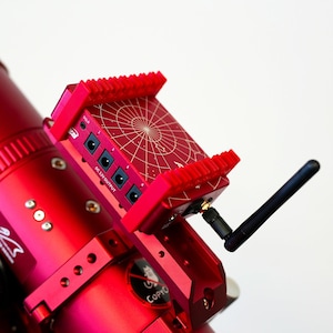 May include: A red telescope with a red power supply unit attached to it. The power supply unit has a star chart on the top and a DC 12V output with four ports. The telescope has a black antenna attached to it.