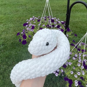 Peut inclure: Un jouet serpent en crochet blanc et moelleux avec un œil noir. Le serpent est fait d'un matériau doux et texturé. L'arrière-plan comprend des paniers suspendus de fleurs violettes et de l'herbe verte.