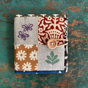 May include: A patchwork journal with a button closure. The cover features various fabric squares with floral, butterfly, and leaf patterns in shades of brown, gray, red, and green. A wooden button and elastic band secure the journal.