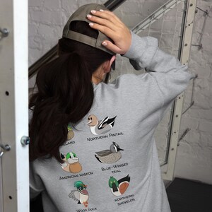 May include: Gray sweatshirt with a design of different types of ducks on the back. The ducks are labeled with their names: Northern Pintail, Blue-Winged Teal, American Wigeon, Wood Duck, and Northern Shoveler.