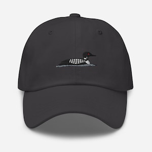 Könnte beinhalten: Dunkelgraue Baseballkappe mit einem gestickten Design eines Eistauchers. Der Eistaucher ist in Schwarz, Weiß und Grau dargestellt, mit einem roten Auge. Die Kappe hat einen gebogenen Schirm und einen Knopf oben.