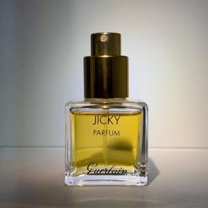 Könnte beinhalten: Ein quadratischer Glasparfümflakon gefüllt mit goldener Flüssigkeit. Der Flakon hat einen goldfarbenen Sprühkopf und Verschluss. Der Text JICKY PARFUM ist in Schwarz auf der Vorderseite aufgedruckt, mit dem Markennamen Guerlain in Schreibschrift.