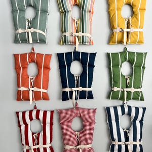 May include: Nine decorative life preservers in various colors and patterns, including stripes and gingham. Each has a central opening and is secured with a white strap and buckle. The colors include green, orange, yellow, and red.