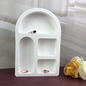 Op de afbeelding: Een witte, gewelfde sieradenorganizer met meerdere compartimenten. De organizer bevat verschillende ringen met diverse edelsteeninstellingen. De achtergrond omvat een transparant gordijn en een boeket gele en witte bloemen.