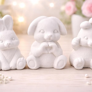 Può includere: Tre statuette di animali in ceramica bianca: un coniglietto, un cucciolo con le mani a forma di cuore e un orso con il pollice in su. Le statuette sono su una superficie chiara, con fiori rosa e verde sullo sfondo.