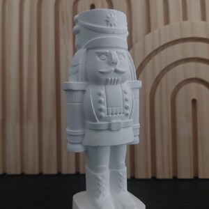 May include: A white, decorative nutcracker figurine. The figurine is in a standing position, with a detailed uniform, hat, and boots. The nutcracker has a mustache and a stern expression. The background is a wooden panel with an arched design.