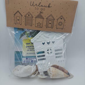 Könnte beinhalten: Ein klarer Plastikbeutel mit einem weißen Strandhütten-Ornament, Muscheln und einer Broschüre. Der Beutel ist mit einem braunen Papieranhänger verschlossen, auf dem die Worte "Urlaub to go" und Illustrationen von Strandhütten aufgedruckt sind.