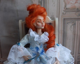 May include: A porcelain doll with red hair wearing a white lace dress with blue accents. The doll is sitting on a white chair with a purple cushion. The doll is wearing blue shoes with white bows.